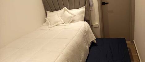 Una cama bien hecha con cabecera gris y ropa de cama blanca, una mesita de noche con lámpara y una puerta a la derecha.