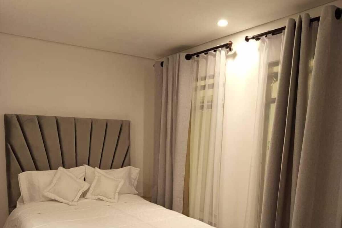A neatly made bed with a headboard, a bedside table, and a window with curtains.