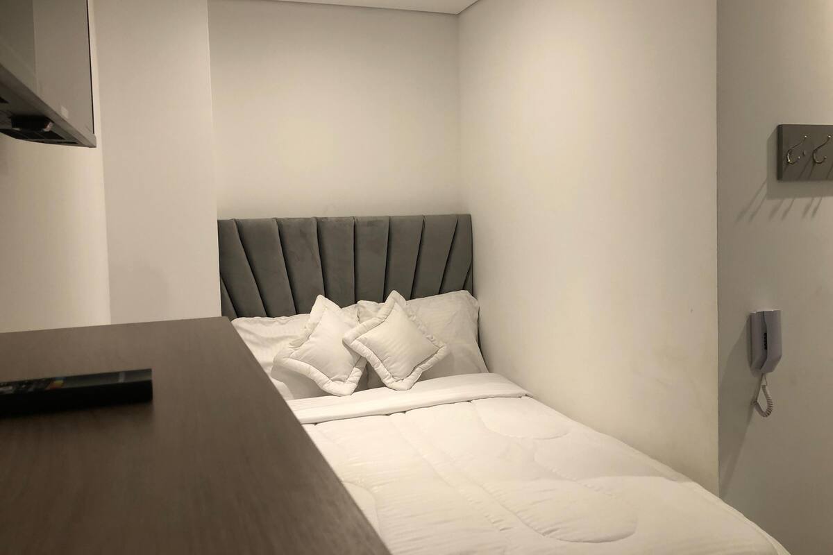 A modern bedroom with a grey upholstered headboard, white bedding, and a bedside telephone.