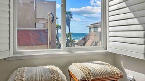 A room with a large window overlooking a beach and ocean, two cushioned seating areas, and a view of a building with a modern design.