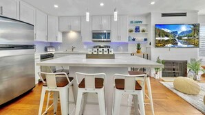A modern kitchen with white cabinetry, a central island with bar stools, a stainless steel refrigerator, and a flat-screen TV mounted above the island.