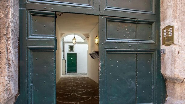 Una puerta grande, de color verde oscuro con superficie texturizada, y una alfombra estampada que conduce a un edificio blanco con una puerta verde y una ventana pequeña.