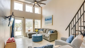 A spacious living room with a large sectional sofa, a round coffee table, and a ceiling fan. There is a staircase with a metal railing leading to an upper level. - 3 Pools and 2 Hot Tubs, Walk to the Beach (Galveston)