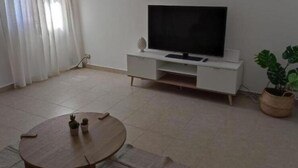 A living room with a flat-screen TV on a white stand, a round wooden table with a remote, a small potted plant, and a large potted plant in the corner. - E&S Apartment (Syracuse)