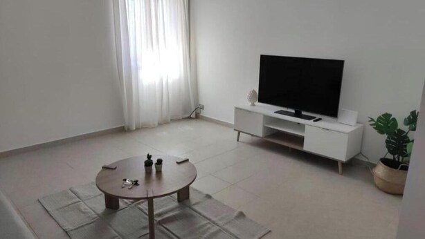 Een woonkamer met een flatscreen televisie op een witte standaard, een ronde salontafel met enkele items en een potplant.