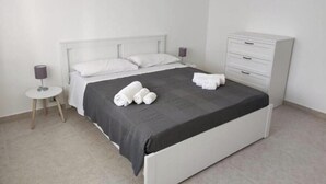 Een netjes opgemaakt bed met witte lakens en een grijze deken, een klein nachtkastje met een lamp, en een witte dressoir met lades.