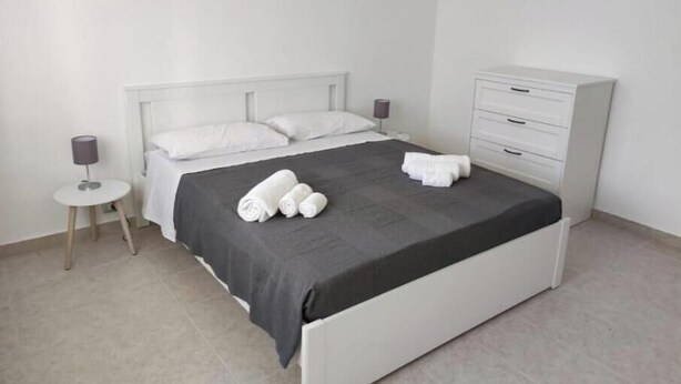 A neatly made bed with white linens and a grey bedspread, a small bedside table with a lamp, and a white dresser with drawers. - E&S Apartment (Syracuse)