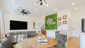Un salon moderne avec une table à manger au plateau en verre, un canapé gris et un mur vert orné d’un néon indiquant « Austin Texas ».