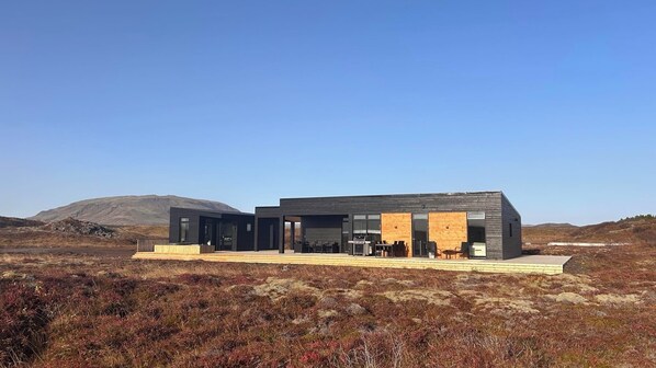 A modern, two-story building with a flat roof, large windows, and a covered outdoor seating area with tables and chairs. - A peaceful, private luxury retreat w/hot tub in the heart of the Golden Circle (Grímsnes- og Grafningshreppur)