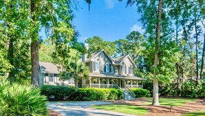Une grande maison à deux étages, dotée d’un porche et entourée d’une végétation luxuriante et d’arbres.