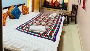 Habitación de hotel con cama, ventilador de techo, unidad de aire acondicionado, ventana con cortinas coloridas y una pequeña mesa con una silla.