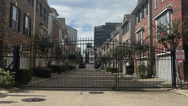 A gated residential street with brick houses, a gated driveway, and a clear sky.
