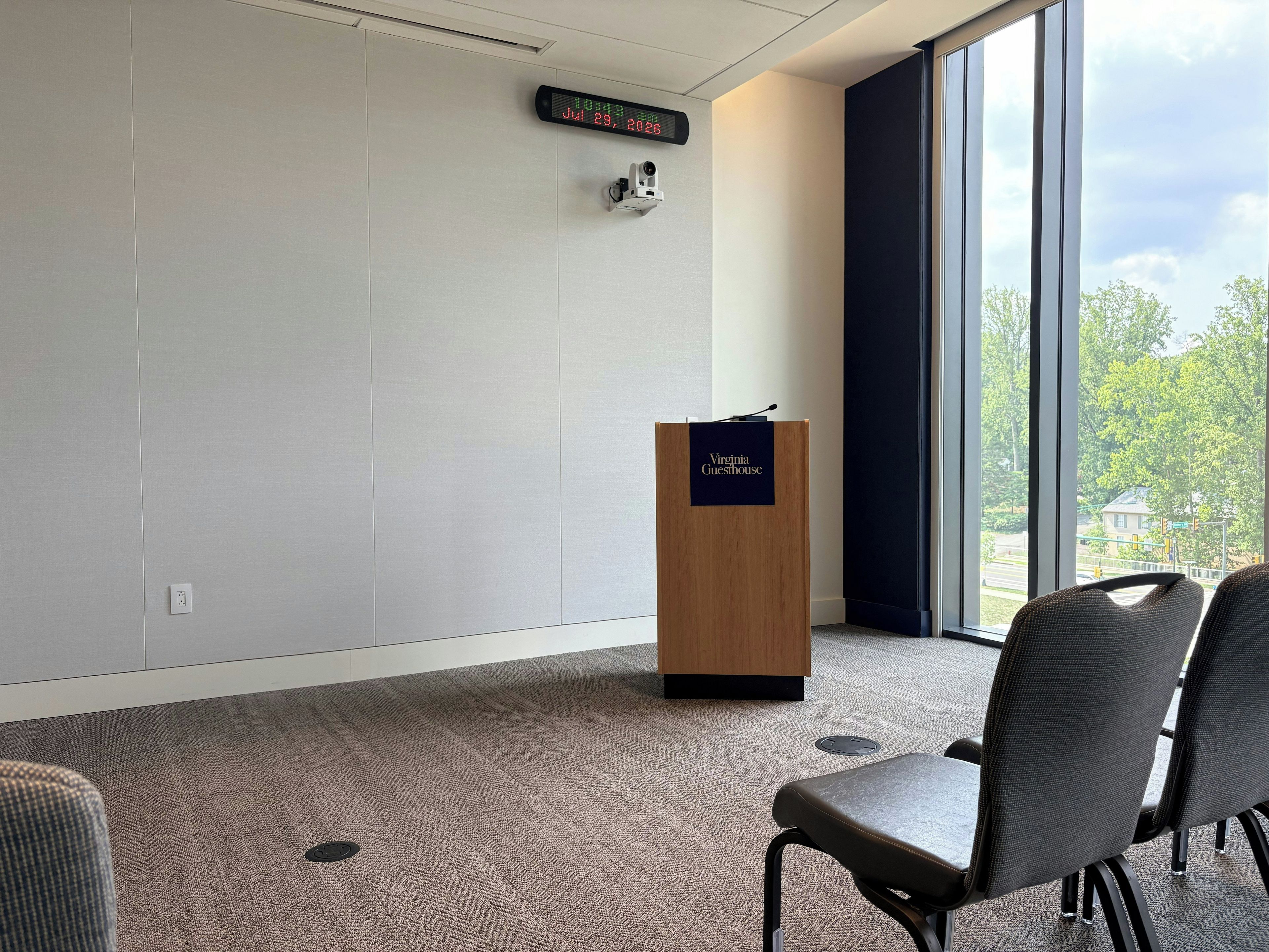 A conference room with a wooden podium labeled 'Virginia Guesthouse,' a digital clock, and a view of trees through large windows.