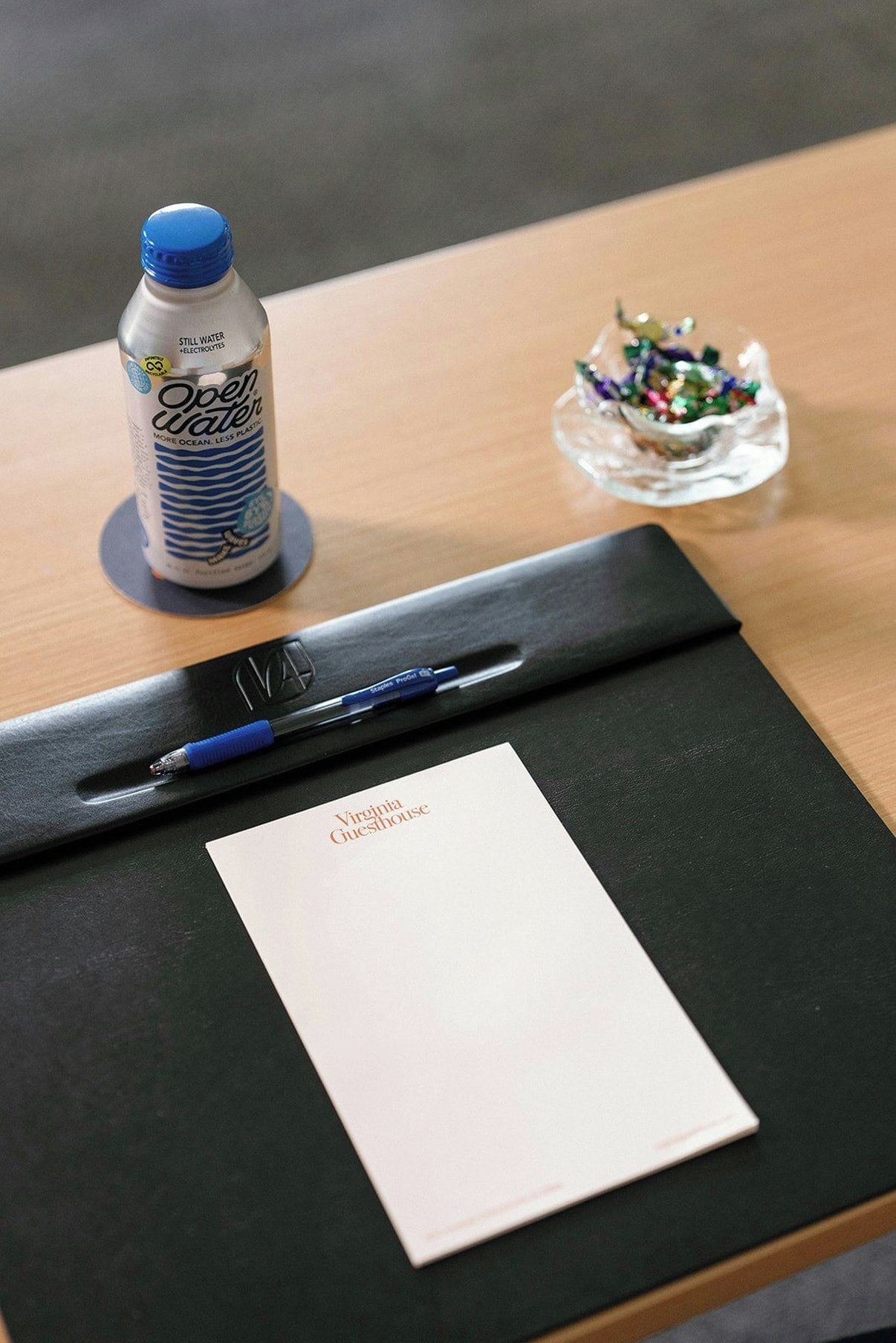 A bottle of still water, a pen, a small glass dish with colorful items, and a piece of paper on a dark surface.