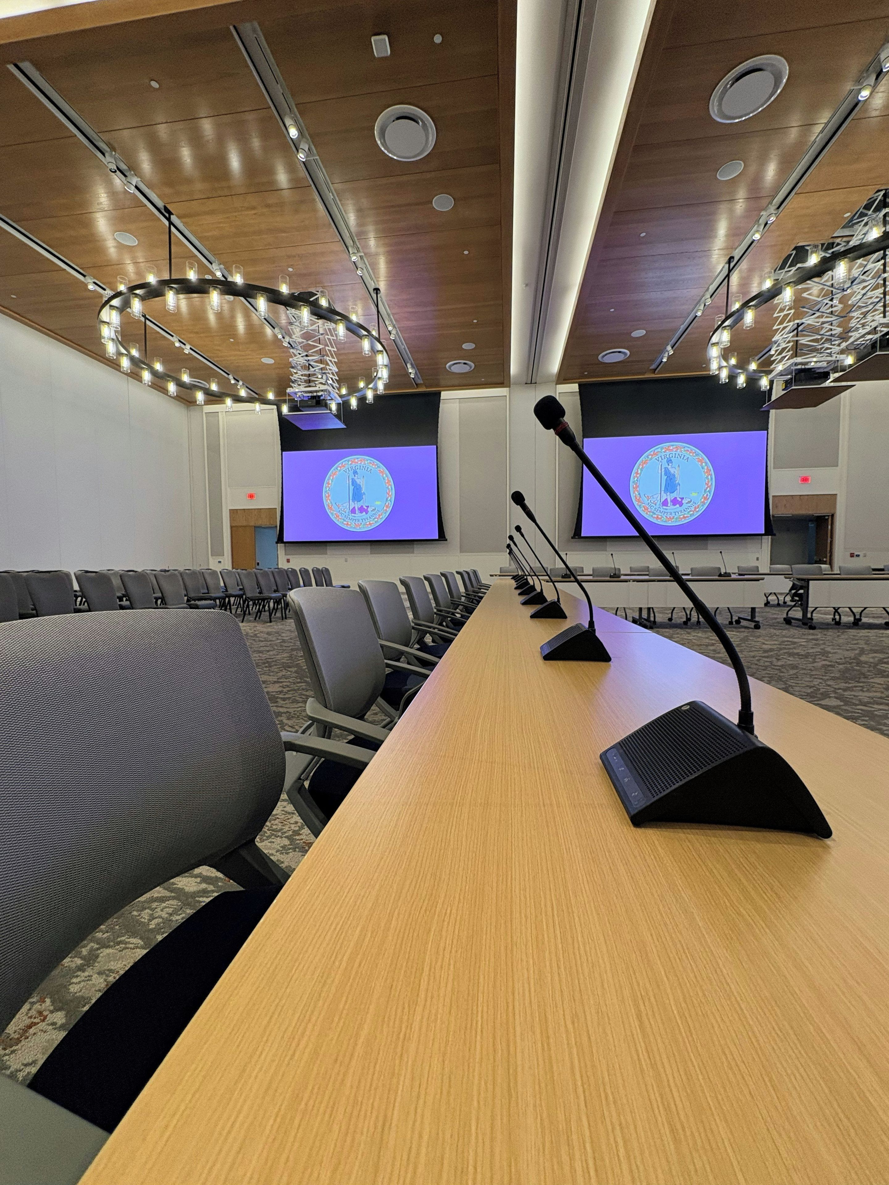 Un salón de conferencias con una larga mesa, varias sillas, un atril con micrófonos y dos pantallas mostrando el emblema del estado de Virginia.
