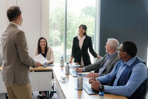 Un salón de conferencias con una larga mesa, sillas y una persona de pie junto a la ventana.