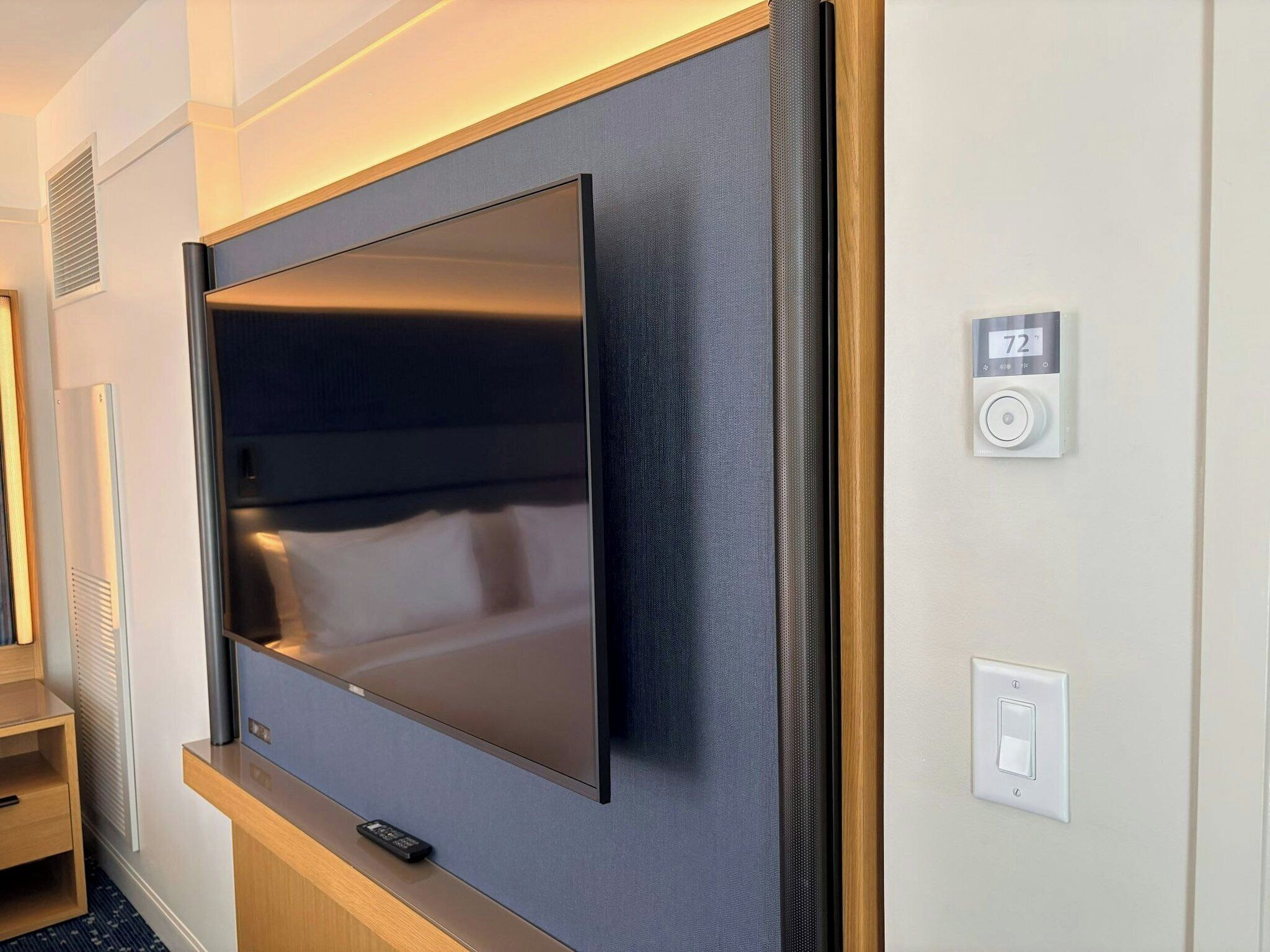 A modern hotel room with a large flat-screen TV mounted on the wall, a remote control on a wooden console, and a wall-mounted thermostat.