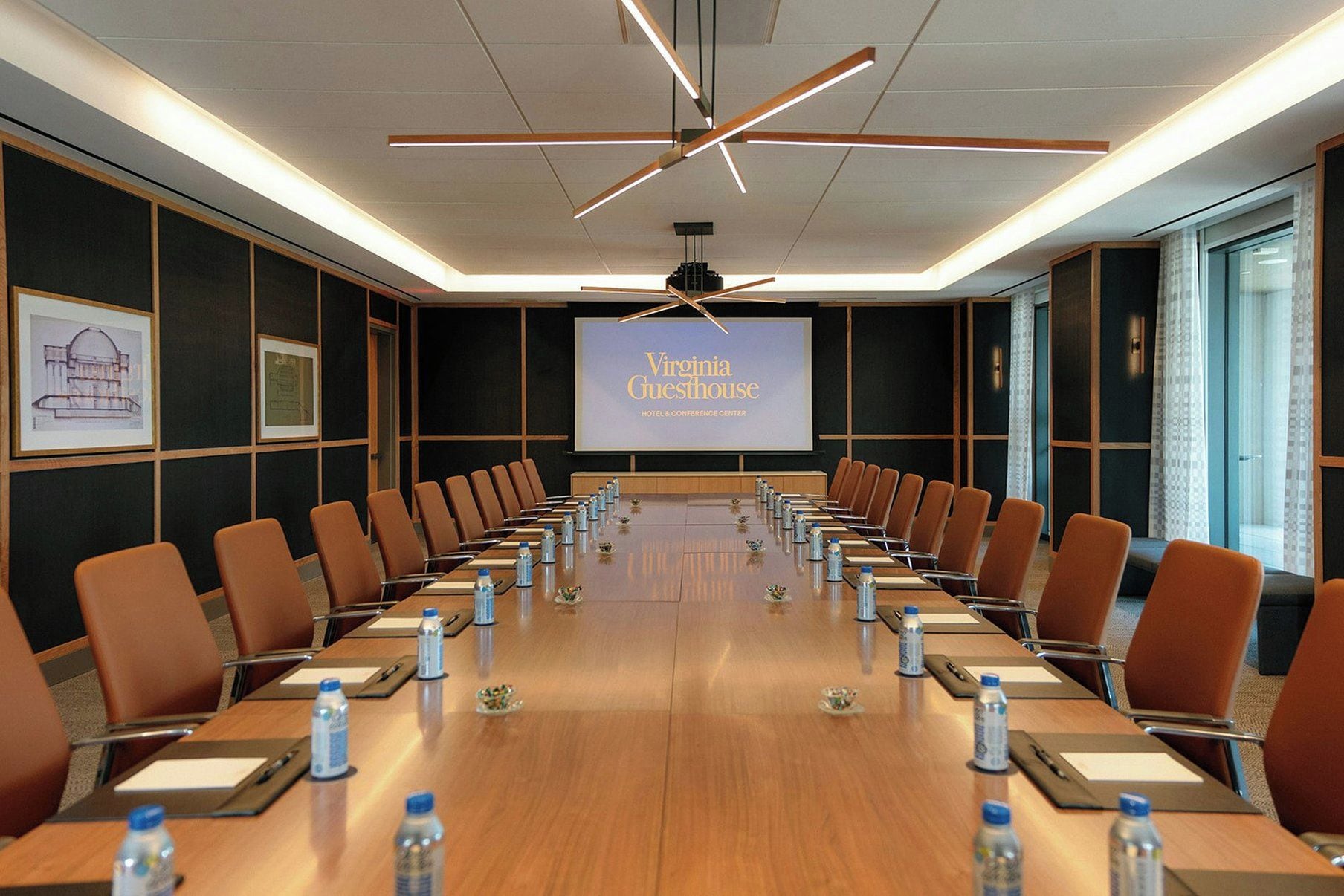 Ein Konferenzraum mit einem langen Tisch, Stühlen, Wasserflaschen und einer Projektionsleinwand, die 'Virginia Guesthouse HOTEL & CONFERENCE CENTER' zeigt.