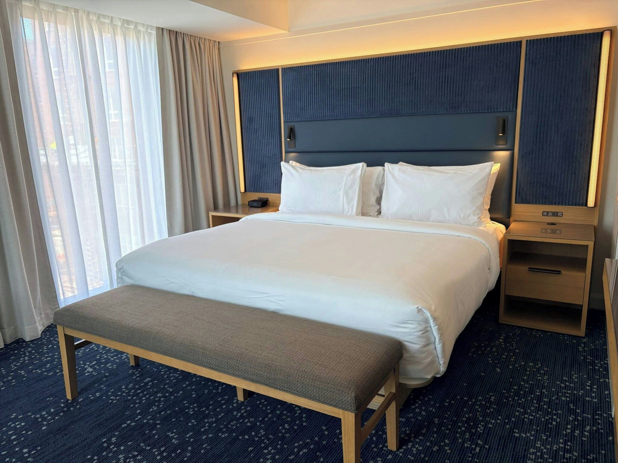 Una habitación de hotel con una cama grande, una mesita de noche, un banco y una mesilla.
