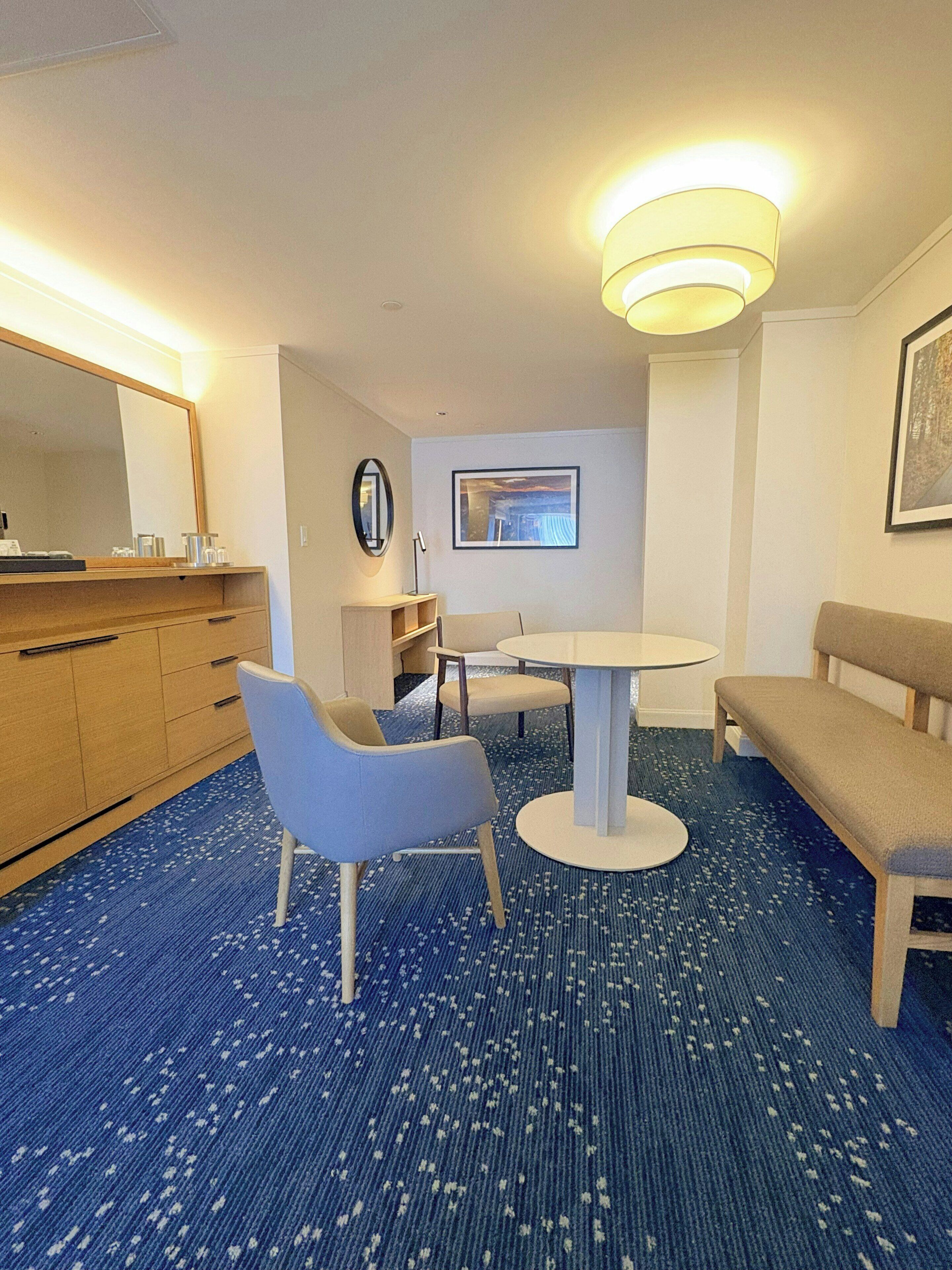 A hotel room with a round table, two chairs, a bench, a dresser, a mirror, and a framed picture on the wall.