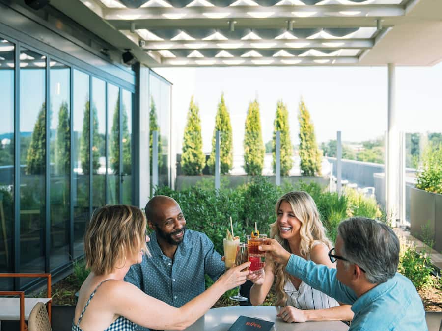 A group of people seated around a table outdoors, with a modern architectural ceiling and large windows offering views of trees and a distant landscape.
