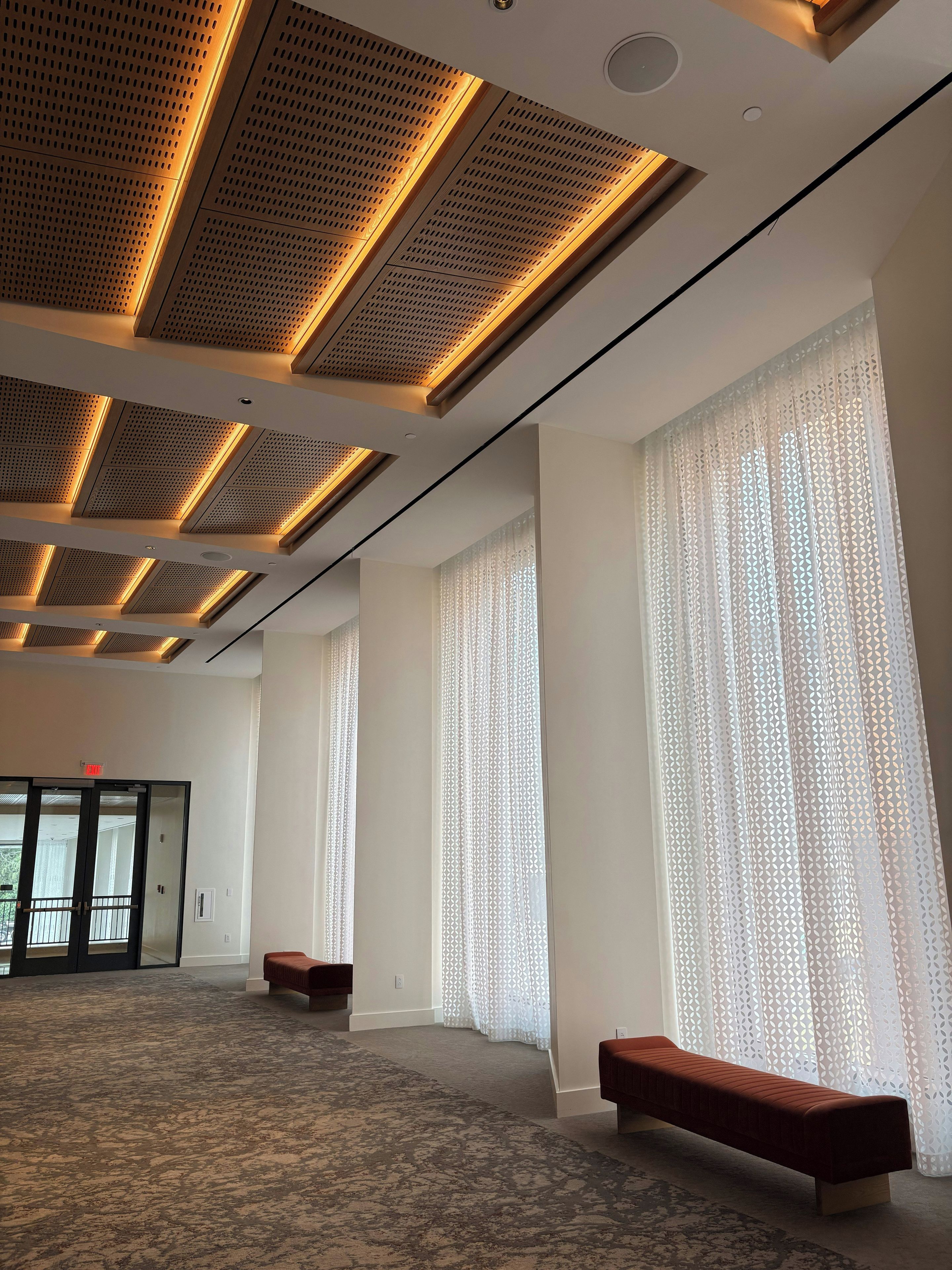 A spacious room with a high ceiling, large windows with patterned curtains, a carpeted floor, and a bench.