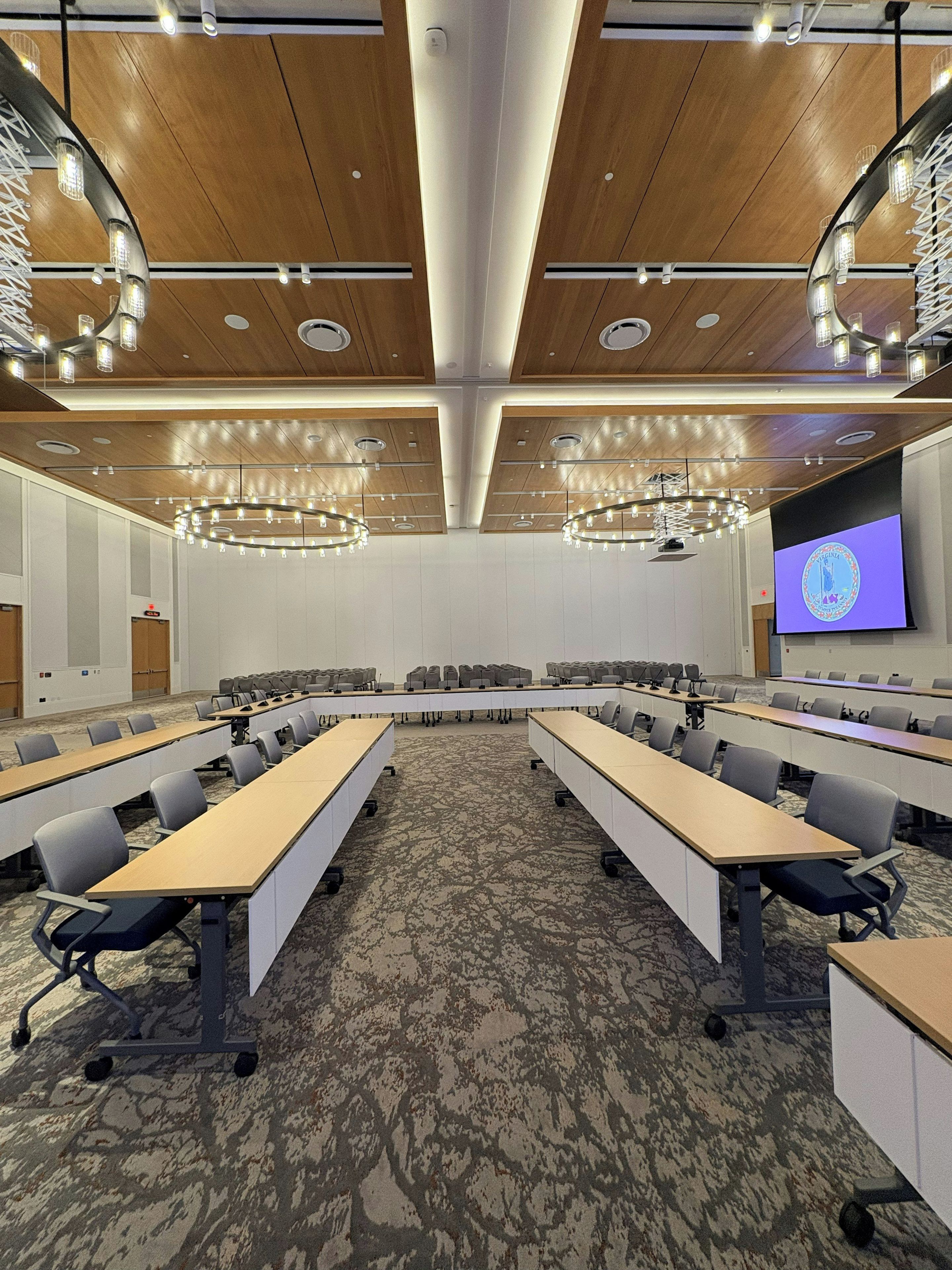 Una sala conferenze con lunghe tavolate, sedie, uno schermo per proiezioni e un soffitto in legno.