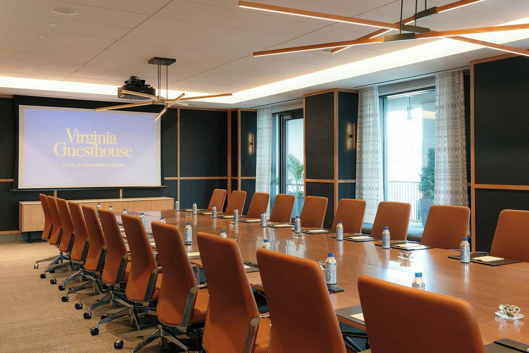 Un salón de conferencias con una larga mesa, varias sillas y una pantalla de proyector mostrando 'Virginia Guesthouse HOTEL & CONFERENCE CENTER'.