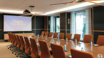 Un salón de conferencias con una larga mesa, varias sillas y una pantalla de proyector mostrando 'Virginia Guesthouse HOTEL & CONFERENCE CENTER'.