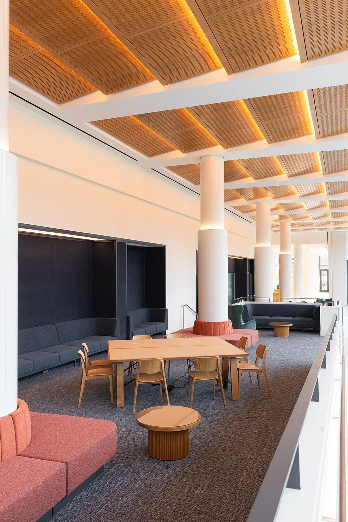 A modern lounge area with a long table, chairs, sofas, and a high ceiling with wooden paneling.