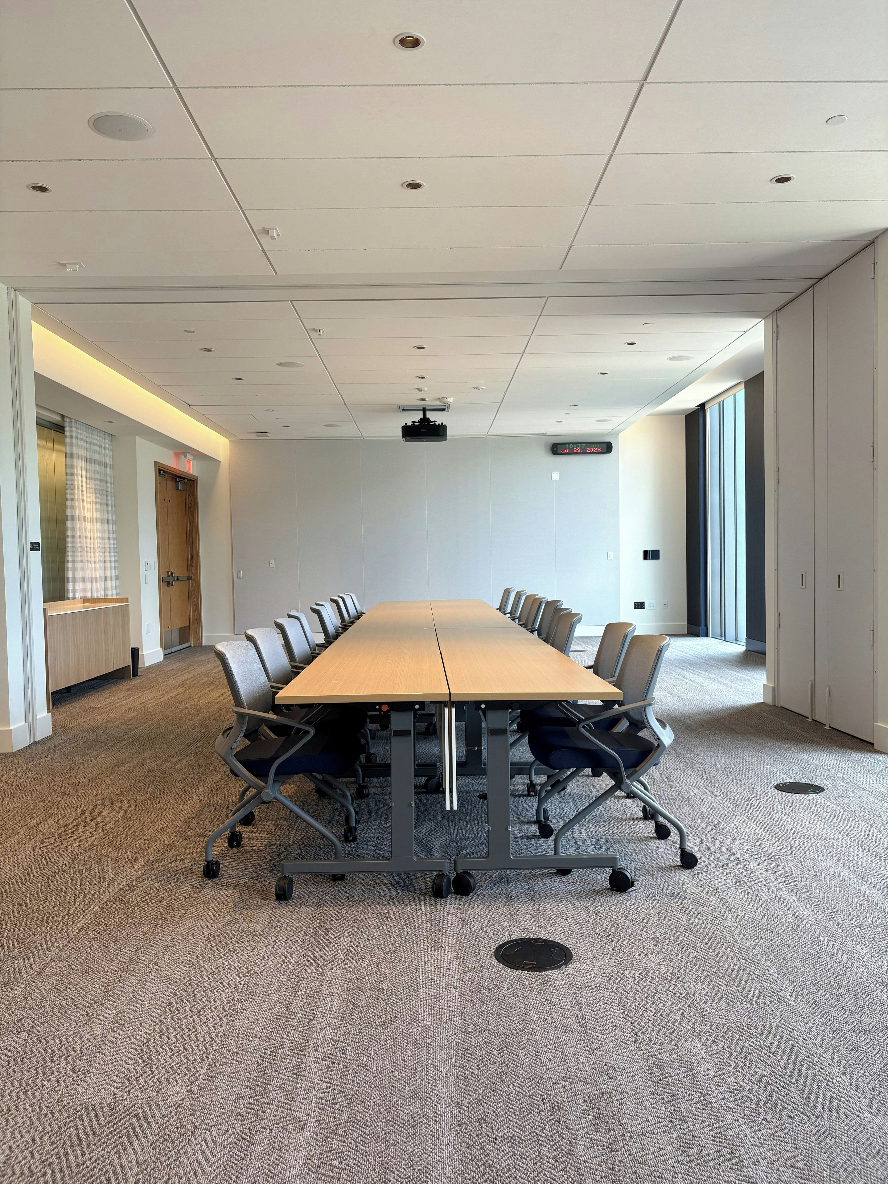 A conference room with a long table, chairs, carpeted floor, ceiling lights, and a projector.