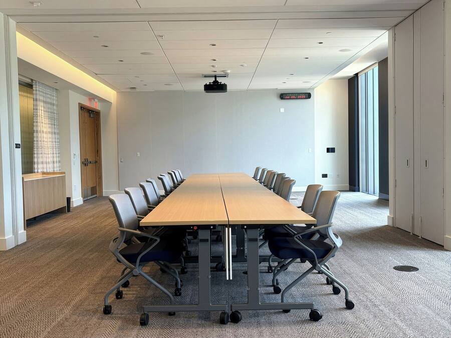 A conference room with a long table, chairs, carpeted floor, ceiling lights, and a projector.