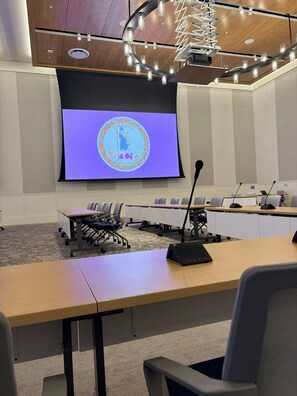 Un salón de conferencias con una pantalla grande mostrando el sello del estado de Virginia, una mesa de madera y sillas de oficina.