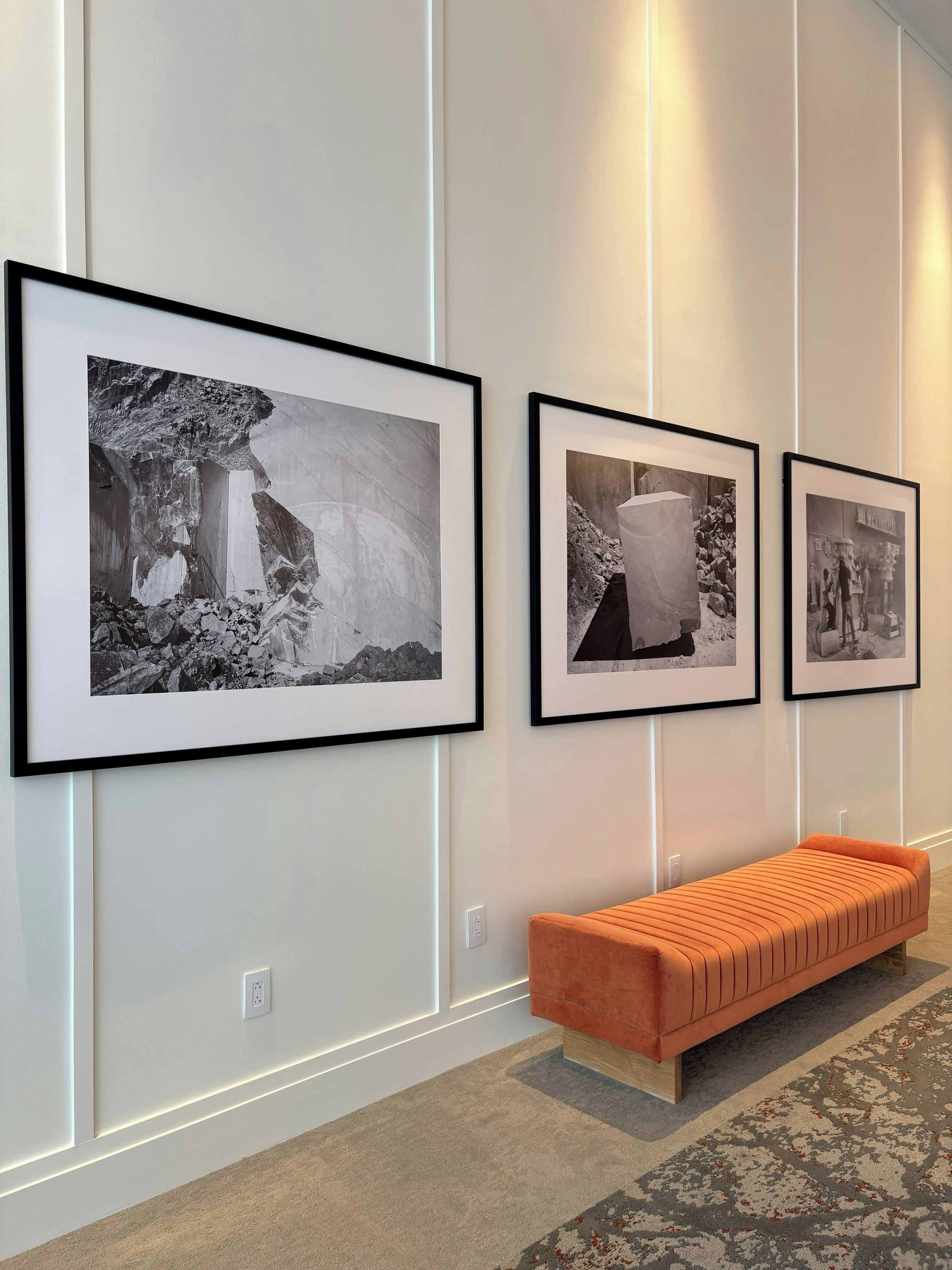 Um corredor com fotografias a preto e branco emolduradas na parede, um banco de madeira e um tapete estampado.