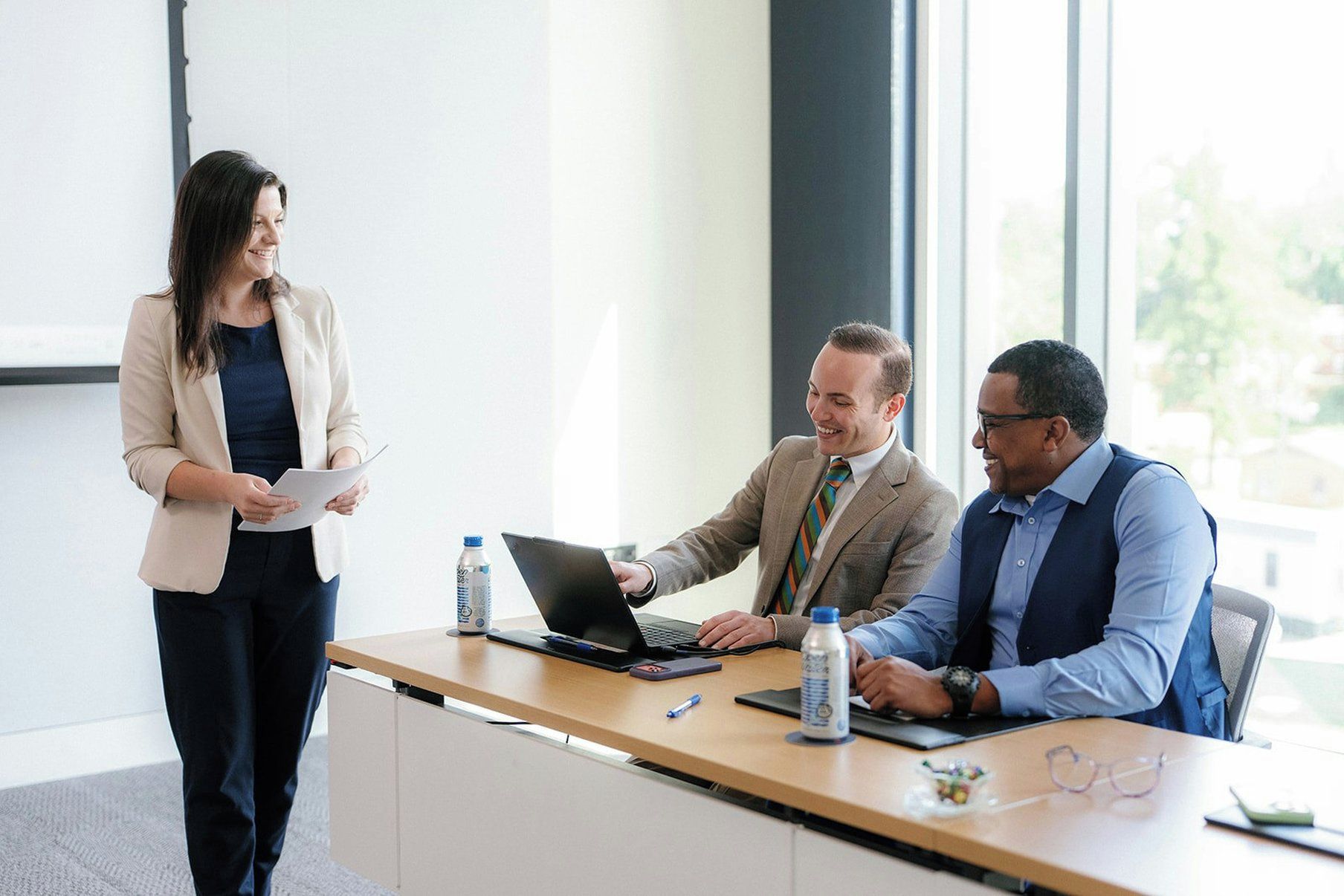 A business meeting in a modern office with a large window, a wooden desk, and a laptop.