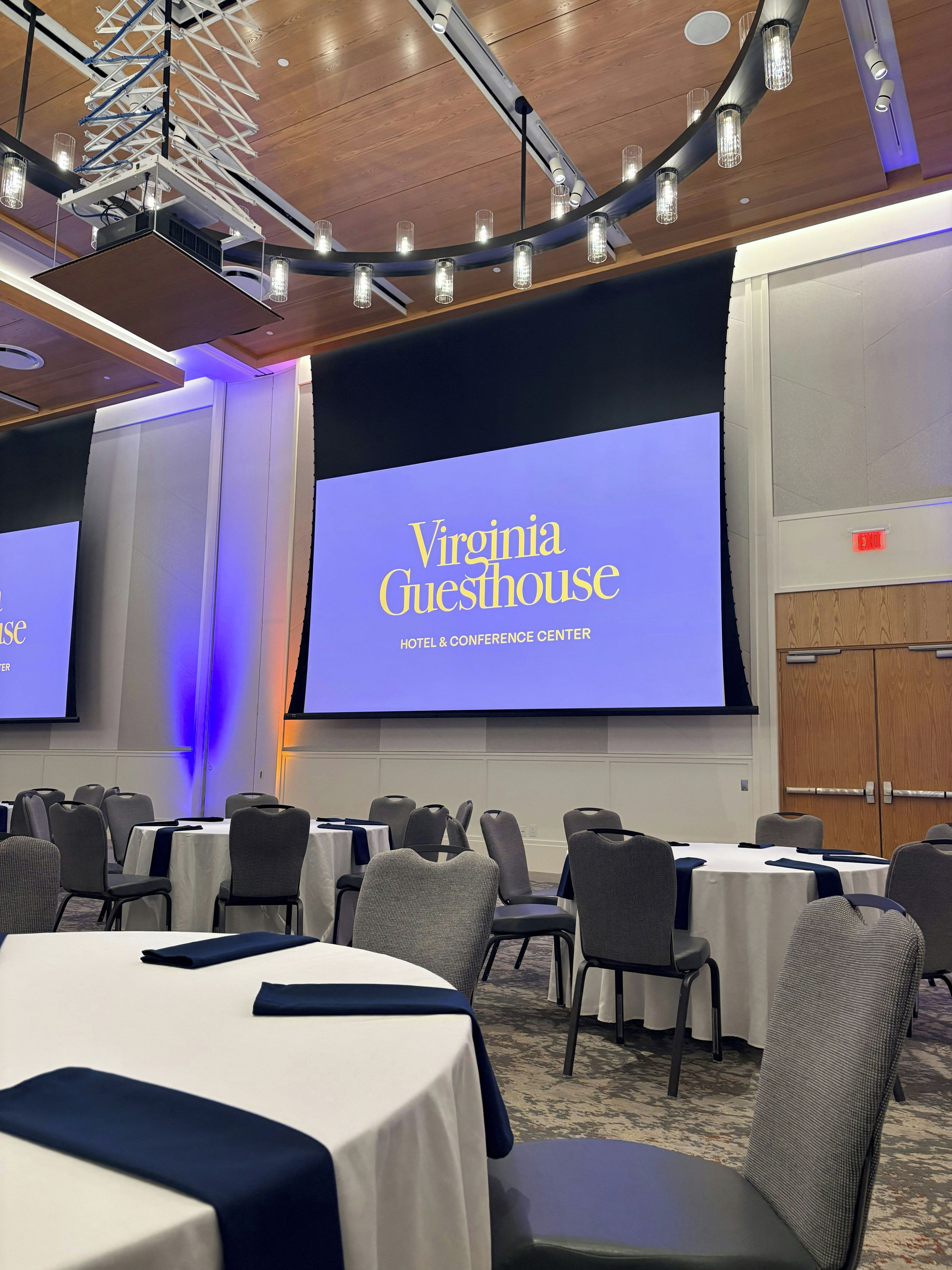 A conference room with tables, chairs, and a large screen displaying 'Virginia Guesthouse HOTEL & CONFERENCE CENTER.'