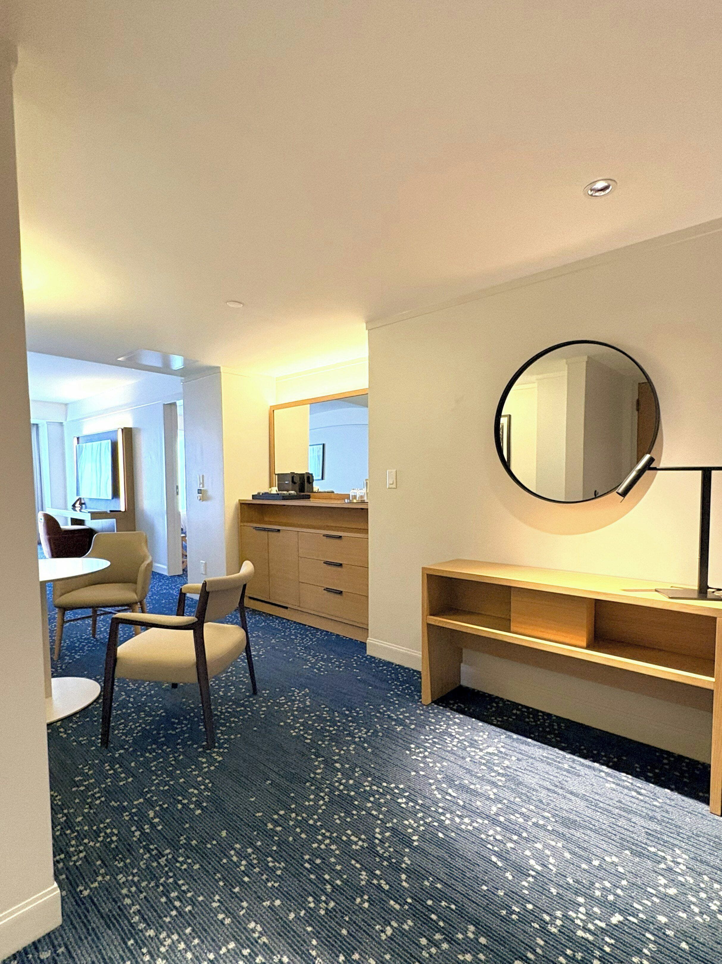A hotel room with a round mirror, a desk, a chair, a small table, and a carpeted floor.