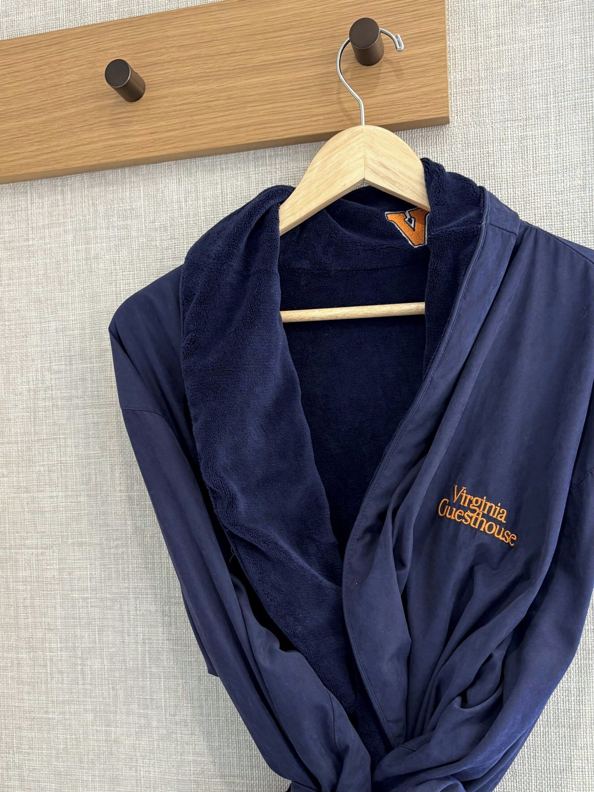 A navy blue robe with 'Virginia Guesthouse' embroidered on it, hanging on a wooden hanger against a textured wall.