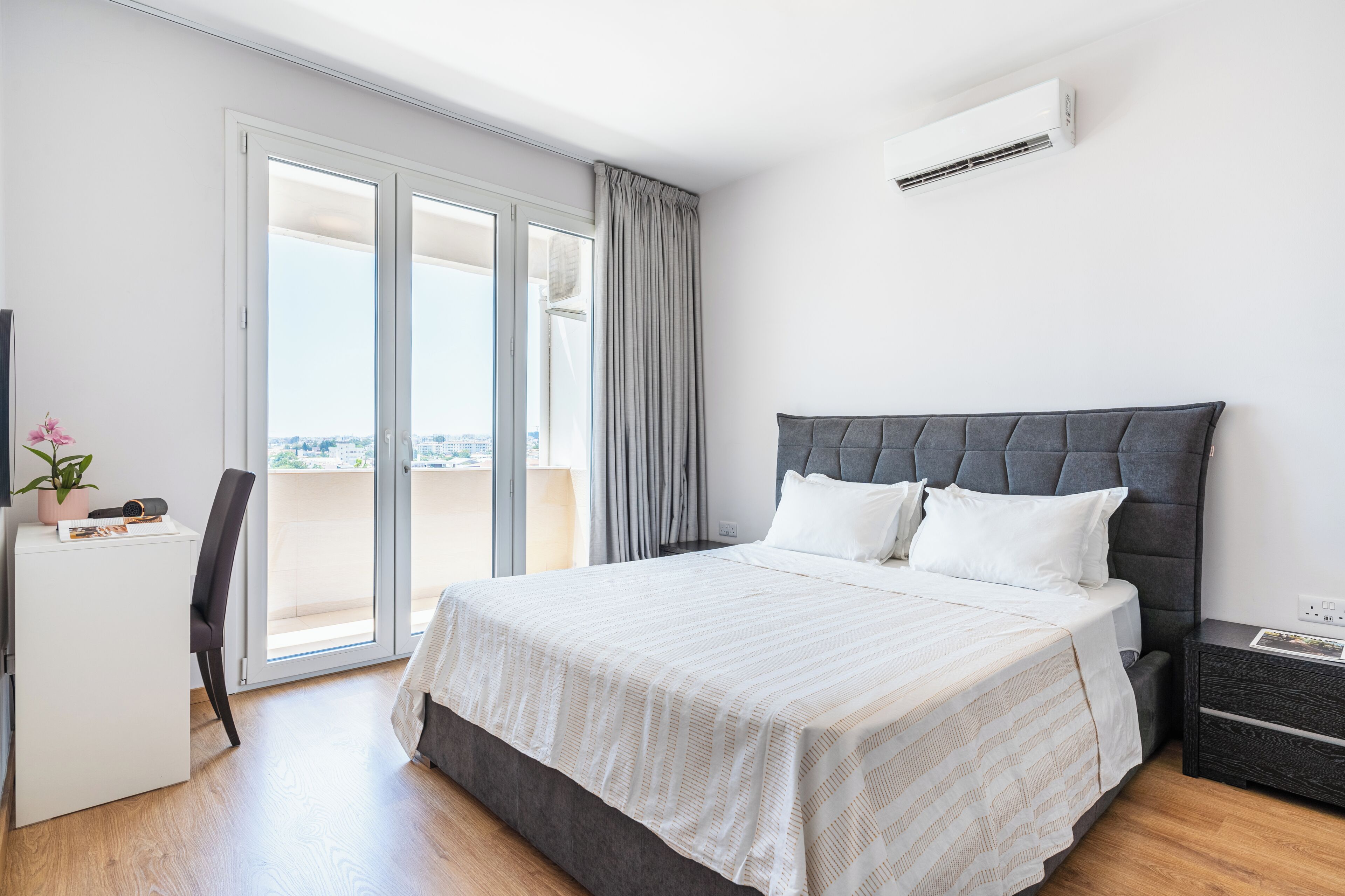 A bedroom with a bed, a desk, a chair, a nightstand, and an air conditioning unit.