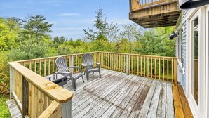 Eine Holzterrasse mit zwei Adirondack-Stühlen, einem Geländer und Blick auf dichtes Grün.
