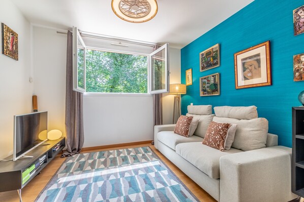 A living room with a sofa, a TV, a patterned rug, a window with curtains, and wall art.