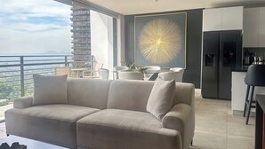 A modern living room with a sofa, a dining area with chairs, a kitchen with a refrigerator, and a large window with a view of buildings and mountains.