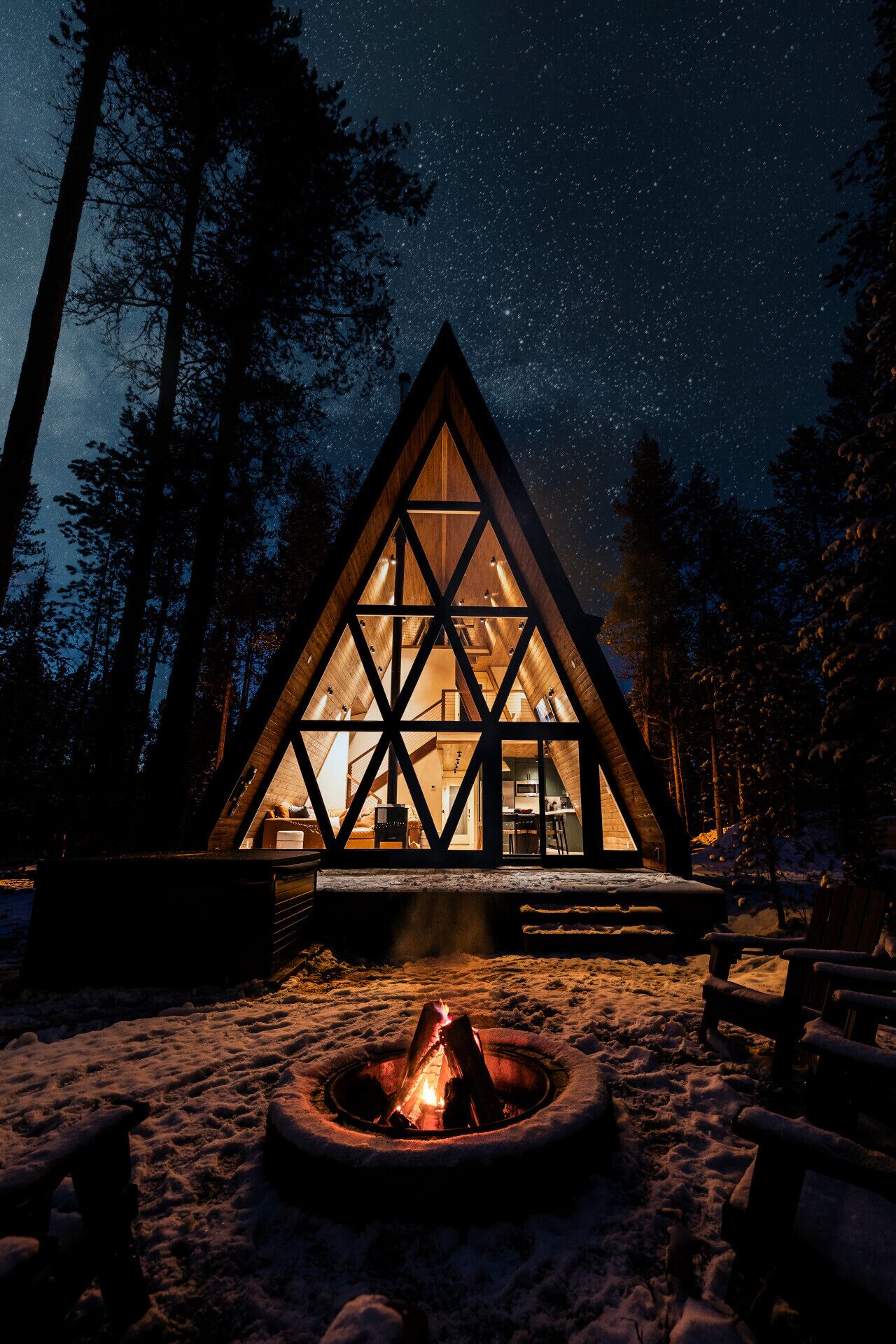 A triangular wooden cabin with a glass facade, surrounded by snow and trees, featuring a fire pit in the foreground.
