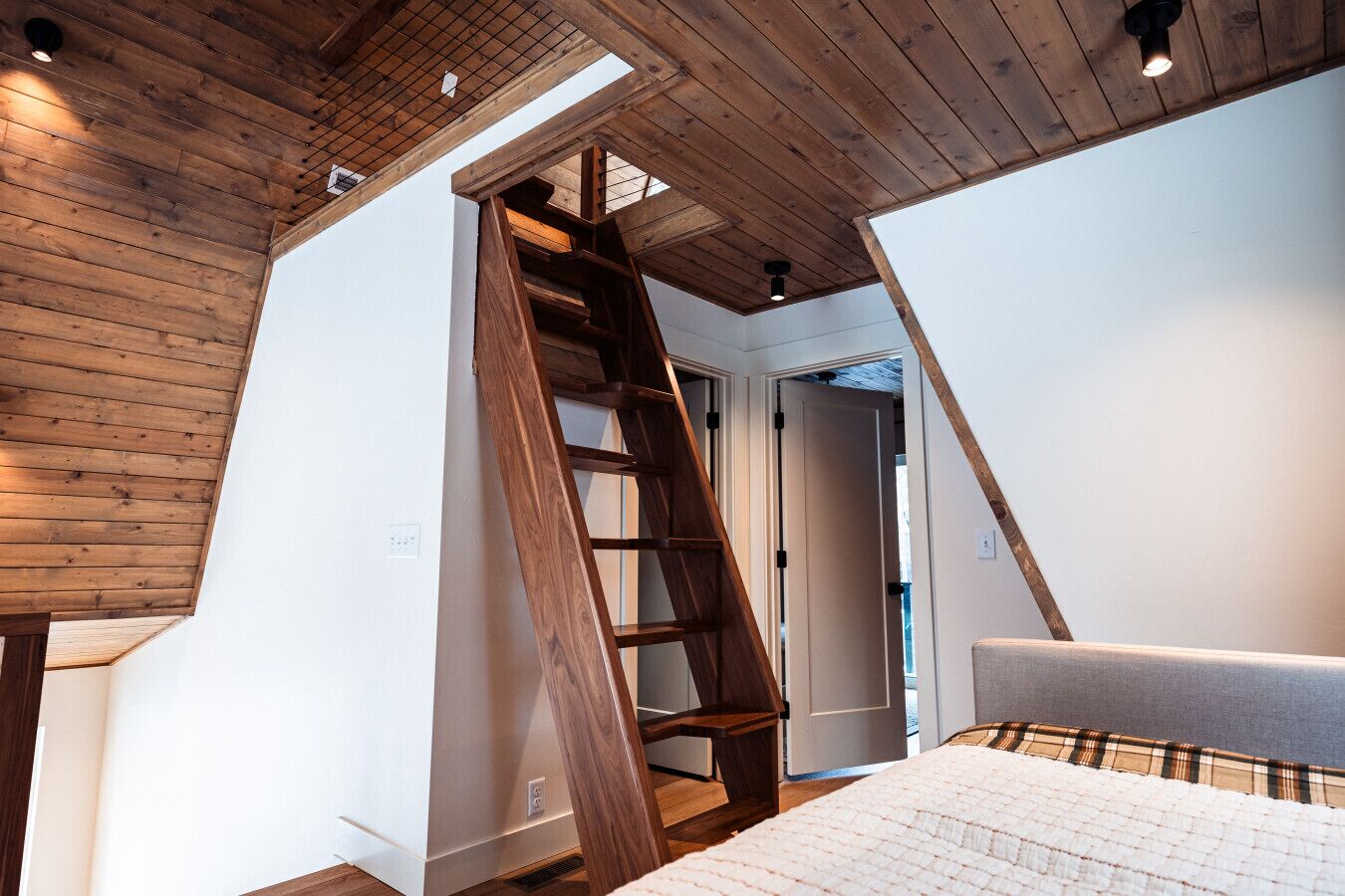 Une chambre avec une échelle en bois menant à une mezzanine aménagée en espace nuit, des murs blancs et un lit recouvert d’une couverture à carreaux.