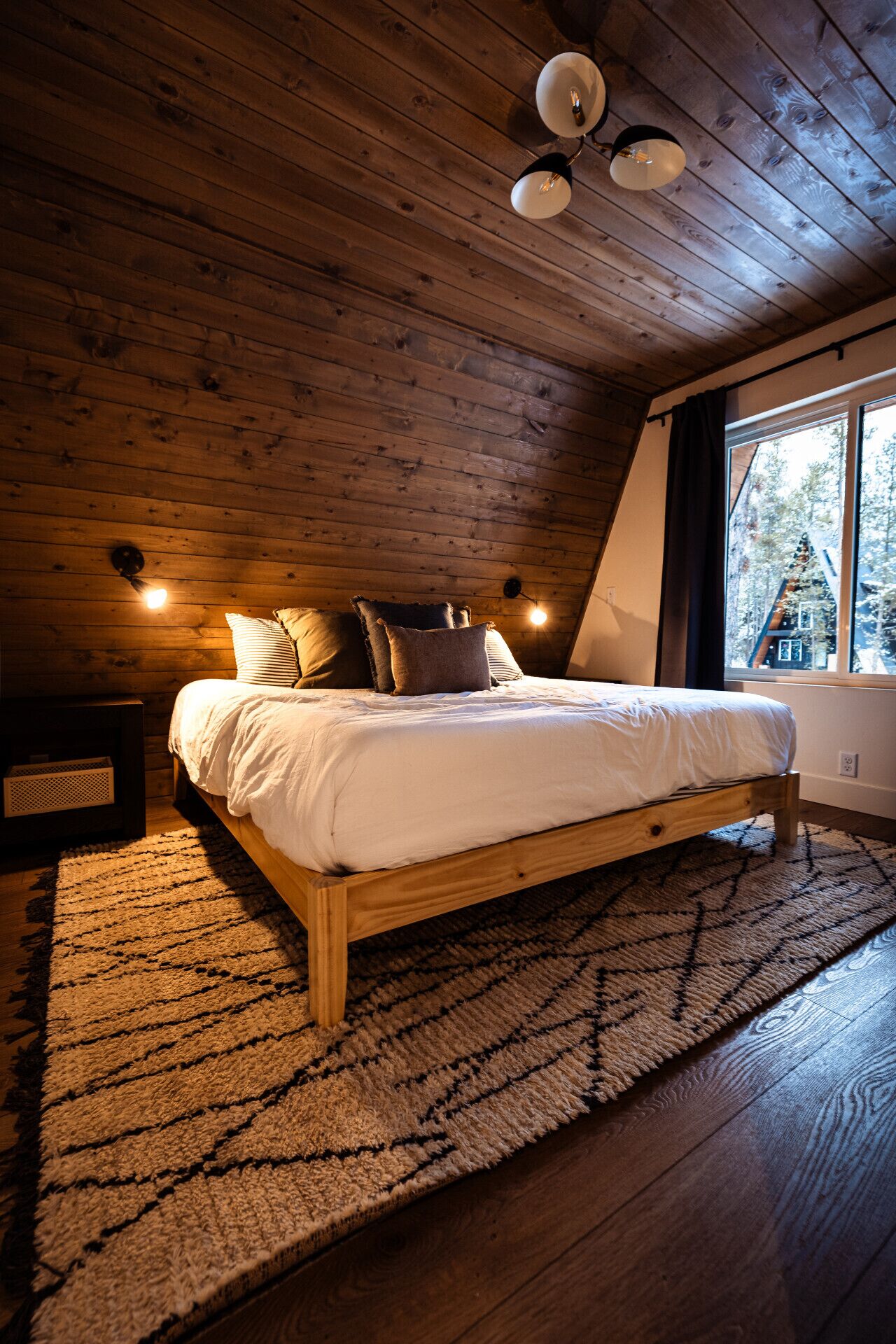 A cozy bedroom with a wooden ceiling, a large bed with white bedding, and a view of a tree through a window.