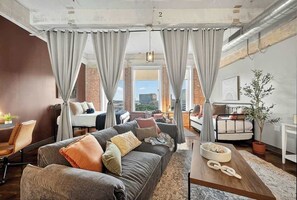 A modern living room with a grey sofa, a wooden coffee table, and a view of the city through large windows. - The Bentley Collection - Loft in Downtown Dallas with Skyline Views (Dallas)