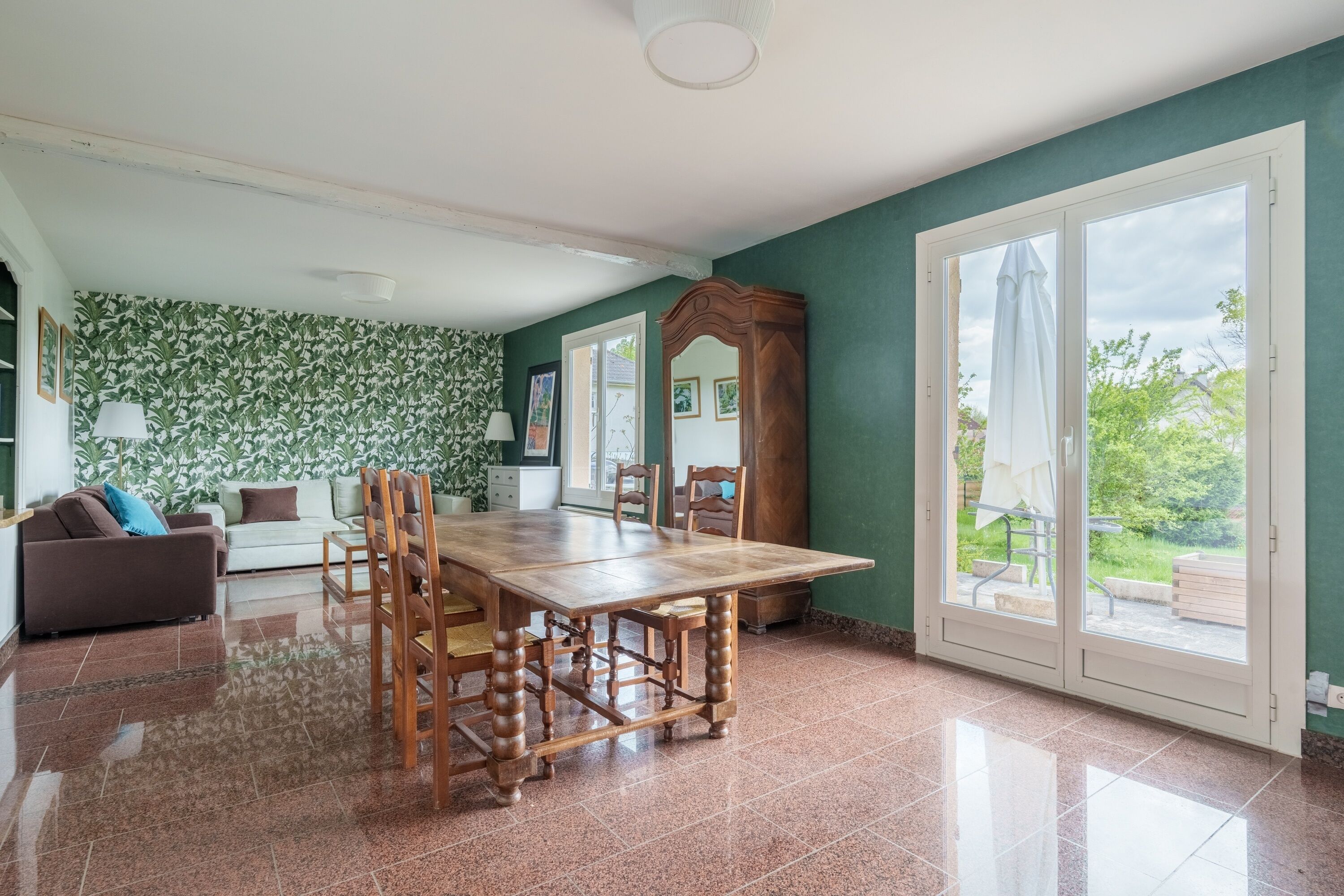 A spacious room with a wooden dining table, a sofa, and a large window with a view of greenery.
