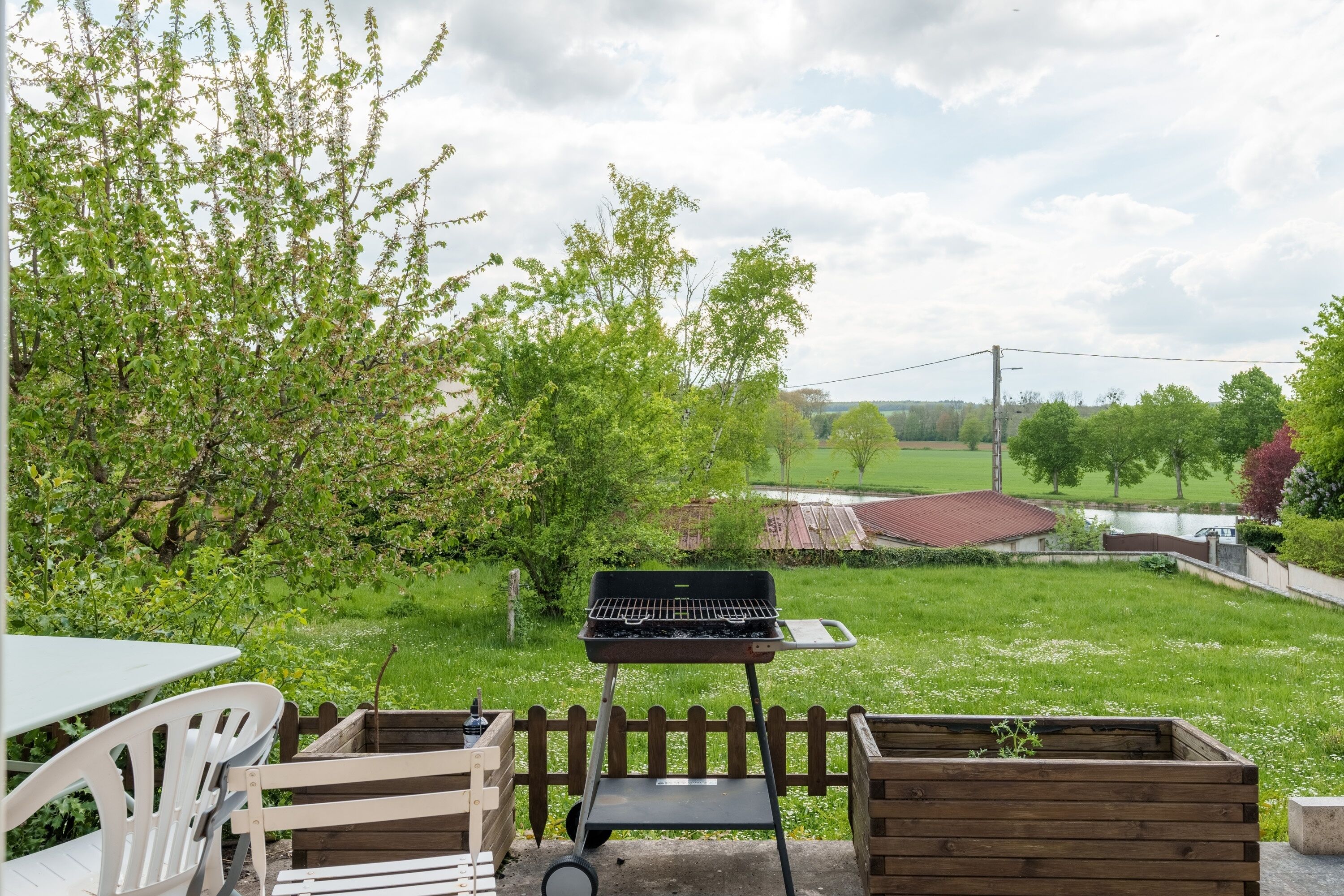 A patio with a grill, a table, and chairs, overlooking a grassy area with trees and a distant building.