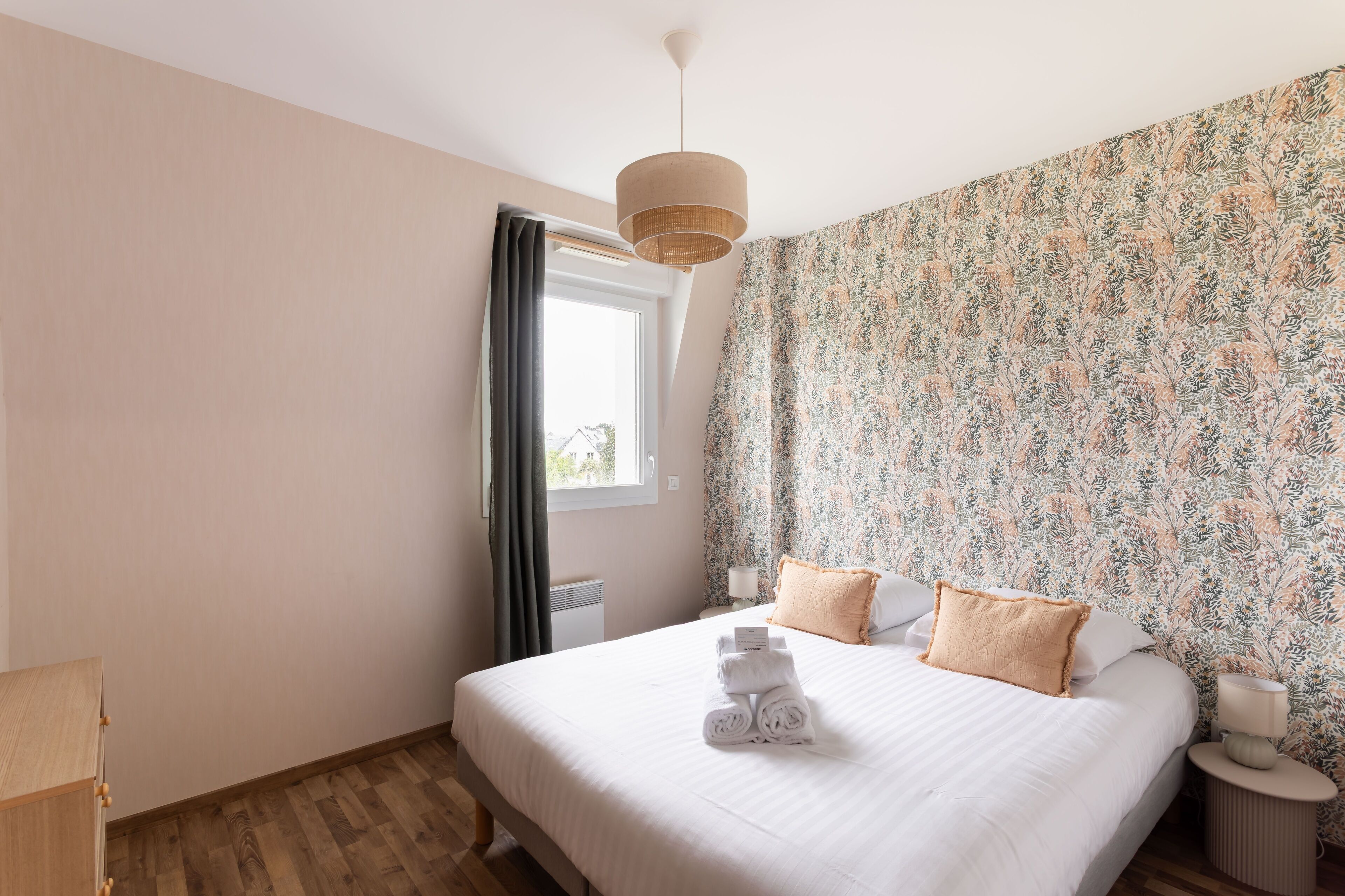 A neatly made bed with rolled towels, a bedside lamp, and a floral-patterned wall.