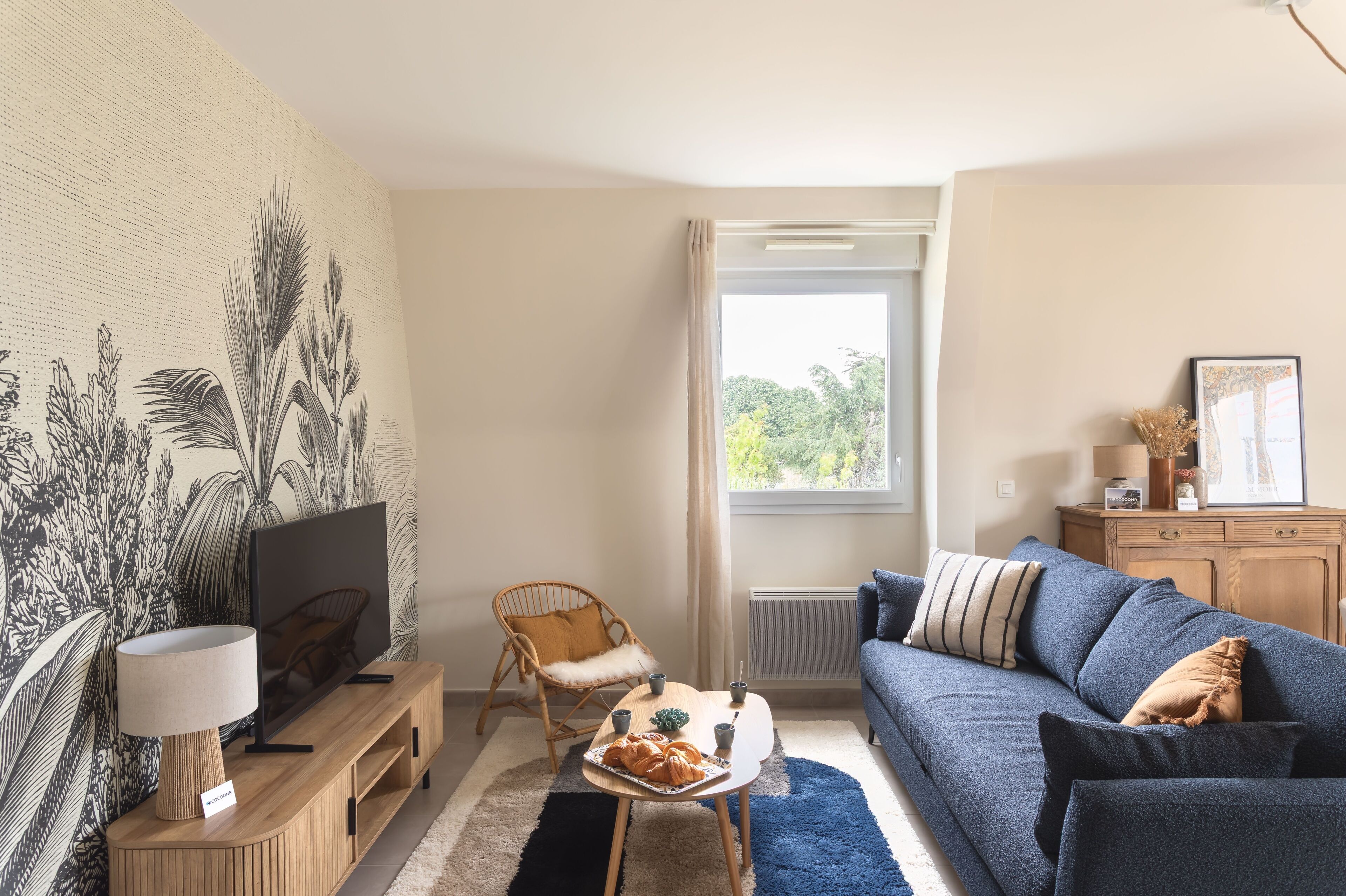 Una sala de estar con televisor, mueble de entretenimiento de madera, sofá azul y un ventanal con vistas a zonas verdes.